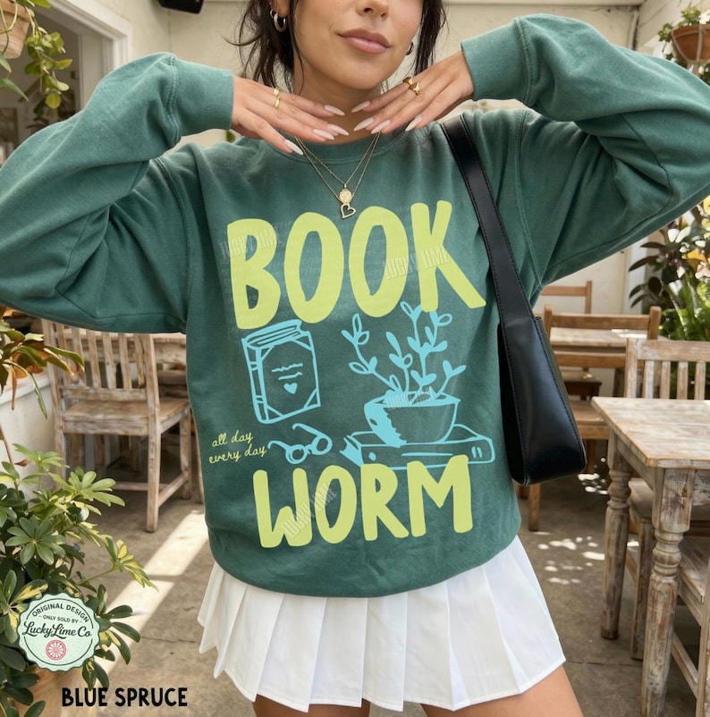 May include: Blue spruce sweatshirt with a graphic that reads "BOOK WORM" in yellow and features a book, plant, and glasses illustration. The text "all day every day" is also included. The sweatshirt has a crew neck and long sleeves.