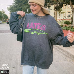 May include: Dark grey sweatshirt featuring the word "LATER" in pink and a green alligator design. The sweatshirt has long sleeves and a crew neck. The text "DARK HEATHER" is printed at the bottom left.