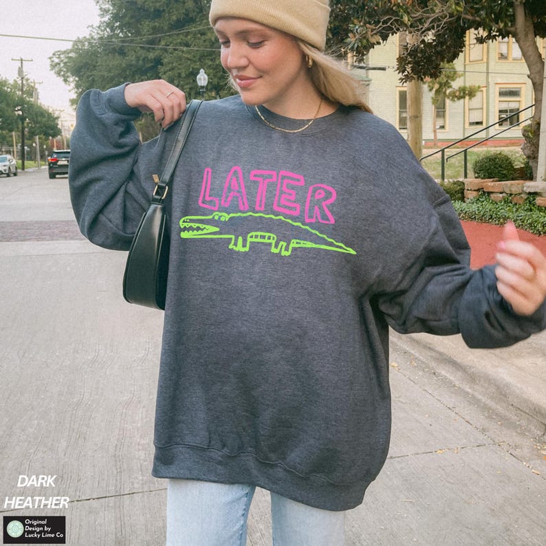 May include: Dark grey sweatshirt featuring the word "LATER" in pink and a green alligator design. The sweatshirt has long sleeves and a crew neck. The text "DARK HEATHER" is printed at the bottom left.