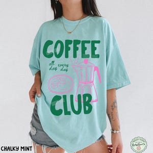 Peut inclure: Un t-shirt vert menthe clair avec le texte vert fonc&eacute; COFFEE CLUB et le texte rose all every day. Un graphique rose repr&eacute;sentant une cafeti&egrave;re italienne et une p&acirc;tisserie est centr&eacute; sur le devant. Le t-shirt a des manches courtes.