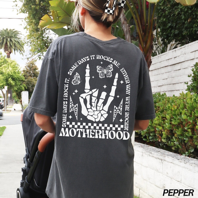 May include: Pepper-colored t-shirt with a white graphic on the back. The graphic includes a skeleton hand making a rock-and-roll sign, butterflies, lightning bolts, and the word "MOTHERHOOD". The text reads "SOME DAYS I ROCK IT. SOME DAYS IT ROCKS ME. EITHER WAY WE'RE ROCKIN".