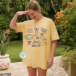 May include: Pale yellow t-shirt with a charming illustration of a teacup, cake, books, butterflies, a bow, flowers, and a jar of lemons. The shirt has the word "BUTTER" at the bottom and a "Lucky Lime Co" logo.