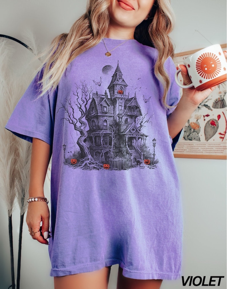 Może przedstawiać: Duży, fioletowy T-shirt z nadrukiem graficznym przedstawiającym straszny nawiedzony dom z pełnią księżyca i nietoperzami. Dom ma dynię z wyciętym uśmiechem w oknie i dynie na ziemi.