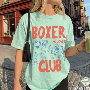 May include: A light mint green short-sleeve t-shirt featuring the red text BOXER mom CLUB and blue line art illustrations of two boxer dogs. A black shoulder bag strap is visible on the left side of the shirt.