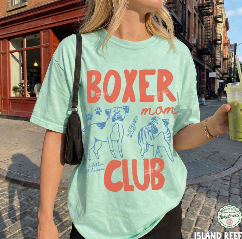 May include: A light mint green short-sleeve t-shirt featuring the red text BOXER mom CLUB and blue line art illustrations of two boxer dogs. A black shoulder bag strap is visible on the left side of the shirt.