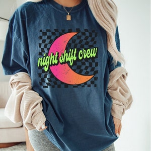 May include: A navy blue t-shirt with a graphic of a crescent moon on a black and white checkered background. The text "night shift crew" is written in green on the moon.