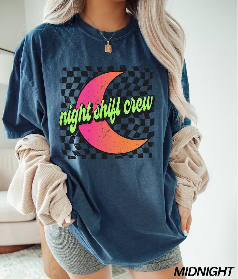 May include: A navy blue t-shirt with a graphic of a crescent moon on a black and white checkered background. The text "night shift crew" is written in green on the moon.