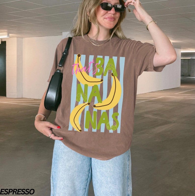 May include: A brown t-shirt with a graphic design featuring yellow bananas, blue stripes, and the text "That's Bananas" in green and pink. The shirt is worn with blue jeans and a black purse.