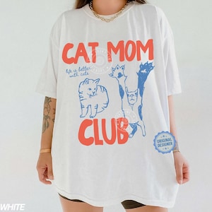 Puede incluir: Camiseta blanca con las palabras "CAT MOM CLUB" en rojo y azul. La camiseta presenta ilustraciones de gatos y la frase "life is better with cats". Una pegatina azul de "Dise&ntilde;ador Original" est&aacute; en la parte inferior derecha.