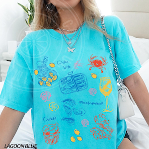 May include: A turquoise blue t-shirt with a variety of colorful illustrations of seafood, lemons, and a glass of wine. The text "Dolce Vita" and "Mediterranean" are printed on the shirt.