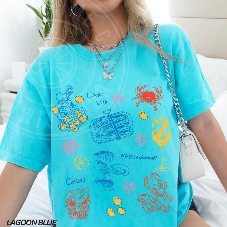 May include: A turquoise blue t-shirt with a variety of colorful illustrations of seafood, lemons, and a glass of wine. The text "Dolce Vita" and "Mediterranean" are printed on the shirt.
