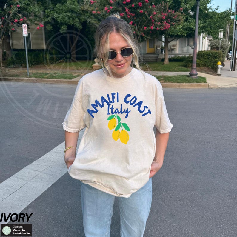 K&ouml;nnte beinhalten: Eine Frau tr&auml;gt ein hellbeiges T-Shirt mit einem blauen Aufdruck, auf dem "Amalfi Coast Italy" steht und eine Gruppe gelber Zitronen abgebildet ist. Das T-Shirt ist oversized und hat eine lockere Passform.