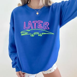 May include: Royal blue sweatshirt featuring the word "LATER" in pink and a green alligator design. The sweatshirt has a crew neck.