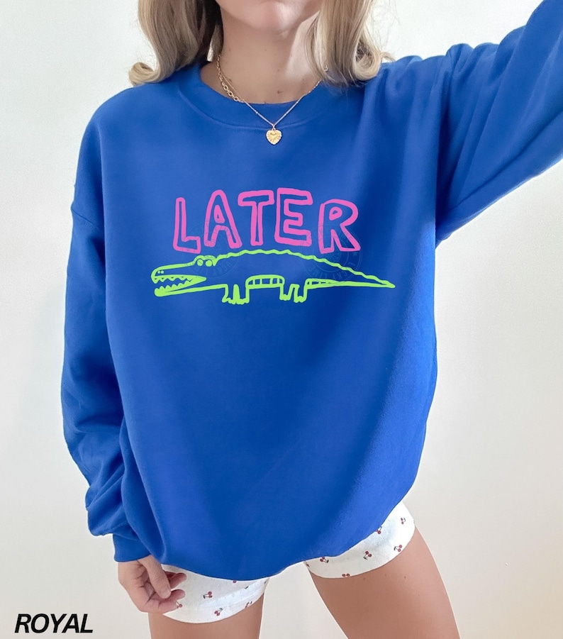 May include: Royal blue sweatshirt featuring the word "LATER" in pink and a green alligator design. The sweatshirt has a crew neck.