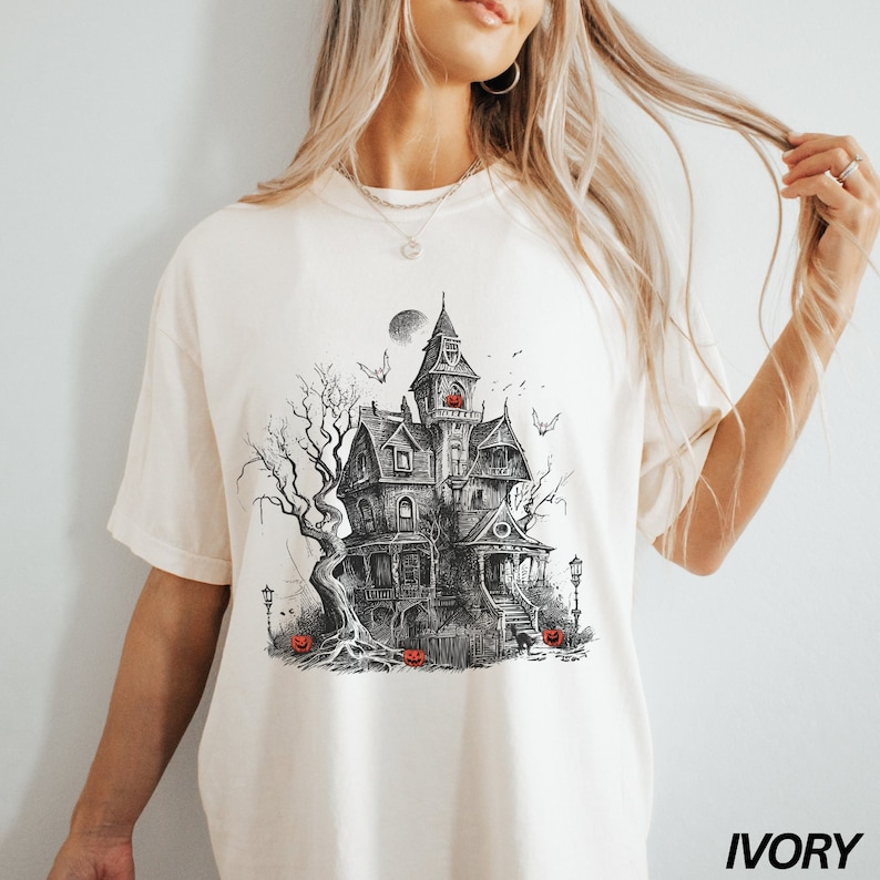 Może przedstawiać: Biały T-shirt z czarno-białą ilustracją nawiedzonego domu z pełnią księżyca i nietoperzami. Dom ma werandę ze schodami i drzewo z korzeniami. Przed domem znajdują się dynie. Tekst "IVORY" znajduje się w prawym dolnym rogu obrazu.