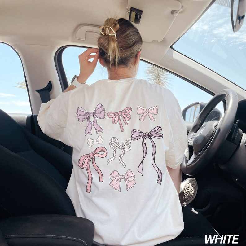 May include: White t-shirt with a pink and purple bow design. The shirt has the word "WHITE" printed on the bottom right corner.