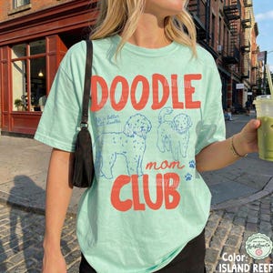 Puede incluir: Camiseta verde claro con las palabras "DOODLE mom CLUB" en rojo. La camiseta presenta ilustraciones azules de dos caniches y la frase "Life is better with doodles". La camiseta es de corte holgado.
