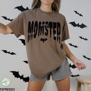 May include: Espresso-colored t-shirt with the word "MOMSTER" in a black, gothic font. The shirt also features a bat graphic and the words "Lucky Lime" in a smaller font. The shirt is part of a Halloween-themed collection.