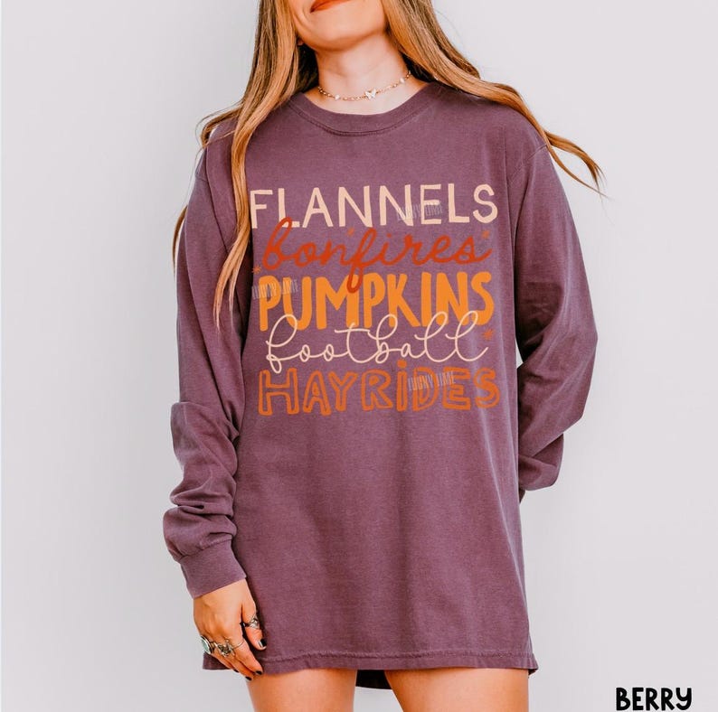May include: A long-sleeve berry-colored crewneck shirt featuring stacked text in various fonts and colors. The graphic displays the words FLANNELS, bonfires, PUMPKINS, football, and HAYRIDES in shades of cream, orange, and light pink.