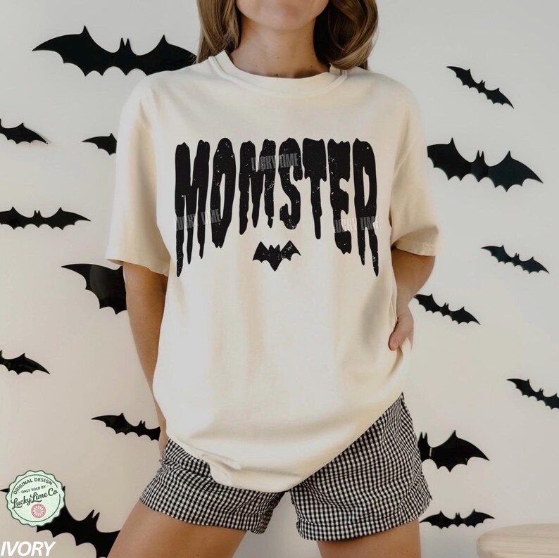 May include: Ivory-colored t-shirt with the word "MOMSTER" in a dripping black font, with a bat graphic. The shirt is paired with black and white checkered shorts. Black bat cutouts decorate the white background.