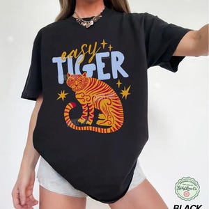 May include: Black t-shirt with a colorful tiger graphic. The design features a tiger with red and yellow stripes, the words "easy TIGER" in blue and yellow, and star accents. The shirt is a casual style.