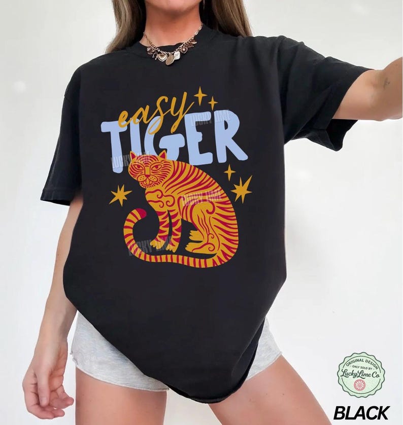 May include: Black t-shirt with a colorful tiger graphic. The design features a tiger with red and yellow stripes, the words "easy TIGER" in blue and yellow, and star accents. The shirt is a casual style.