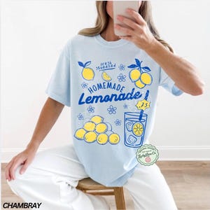 以下が含まれることがあります： 「Homemade Lemonade」のグラフィックがプリントされた水色のTシャツ。レモン、花、レモネードのグラスがデザインされています。「fresh squeezed」の文字も含まれています。ゆったりとしたフィット感のシャツです。