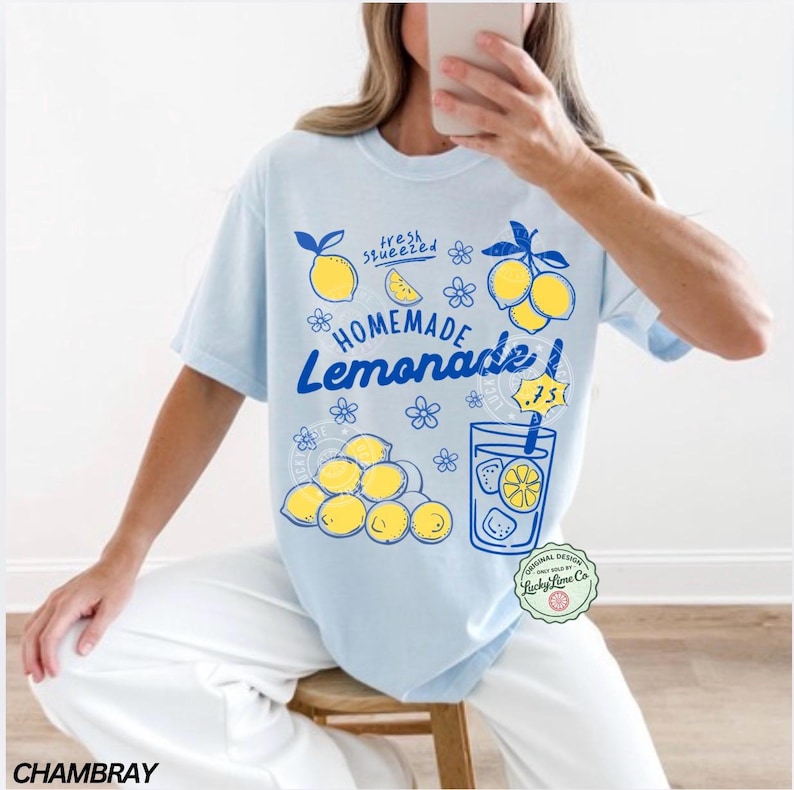 以下が含まれることがあります： 「Homemade Lemonade」のグラフィックがプリントされた水色のTシャツ。レモン、花、レモネードのグラスがデザインされています。「fresh squeezed」の文字も含まれています。ゆったりとしたフィット感のシャツです。