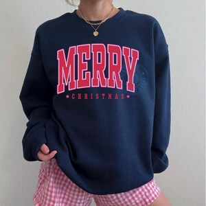 May include: Navy blue sweatshirt with the word "MERRY" in red block letters and "CHRISTMAS" below, with stars. The sweatshirt is paired with pink and white checkered shorts. A gold necklace is visible.