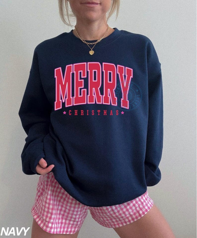 May include: Navy blue sweatshirt with the word "MERRY" in red block letters and "CHRISTMAS" below, with stars. The sweatshirt is paired with pink and white checkered shorts. A gold necklace is visible.