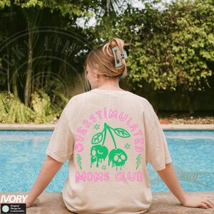 May include: A light beige t-shirt featuring a pink and green graphic of two cherries with skulls on top. The text "Overstimulated Moms Club" is printed in pink below the cherries.