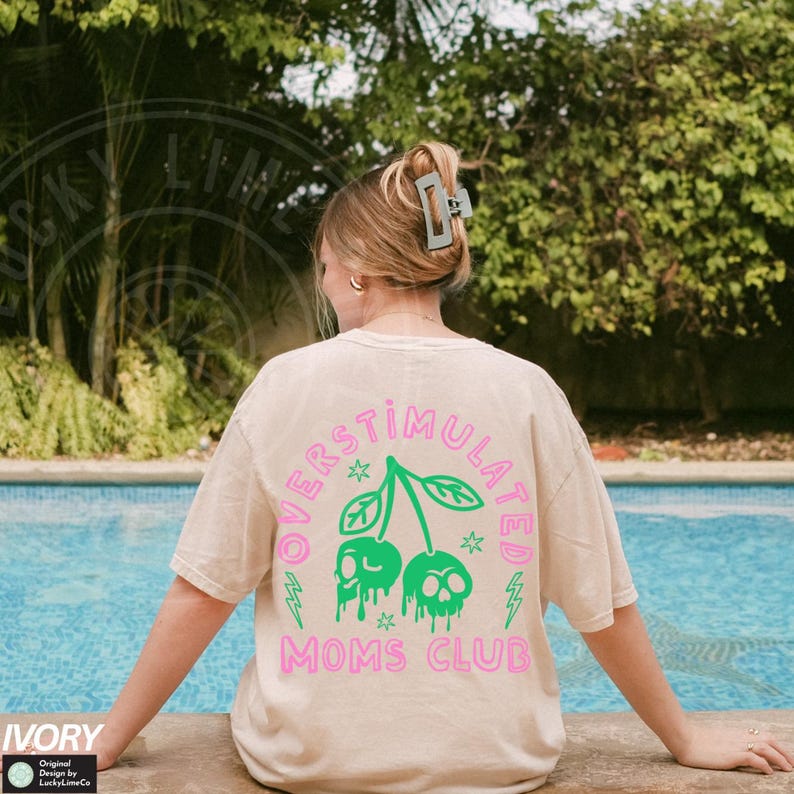 May include: A light beige t-shirt featuring a pink and green graphic of two cherries with skulls on top. The text "Overstimulated Moms Club" is printed in pink below the cherries.