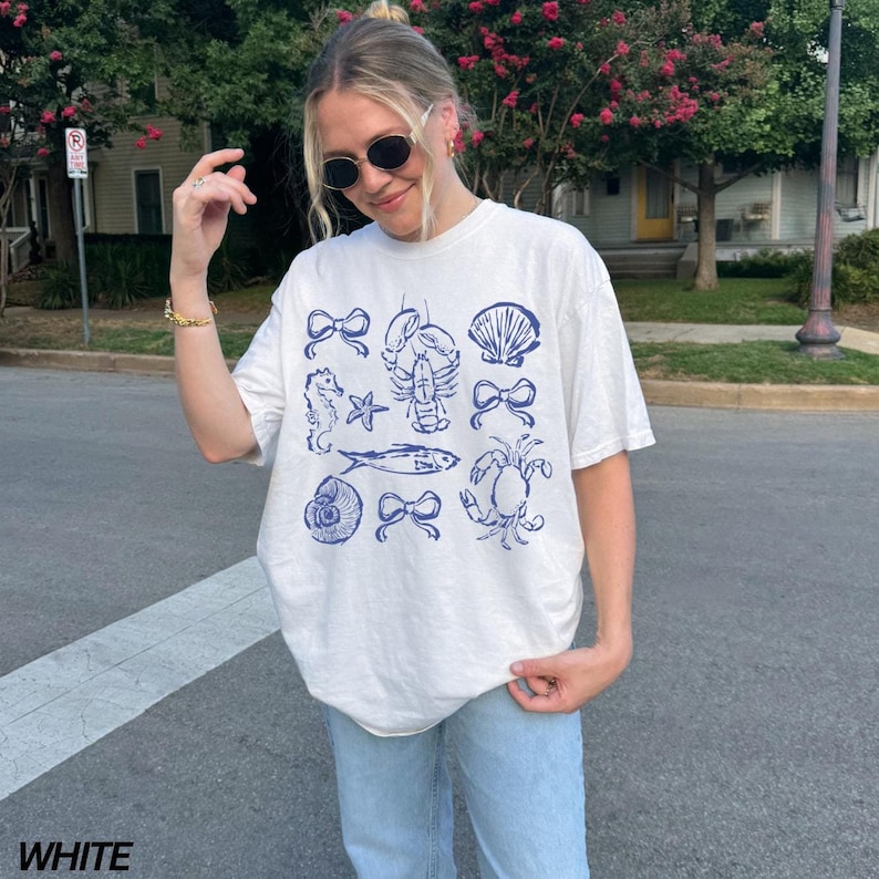 May include: A white t-shirt with a blue graphic print of various sea creatures and bows. The graphic includes a seahorse, starfish, lobster, seashell, fish, crab, and bows.