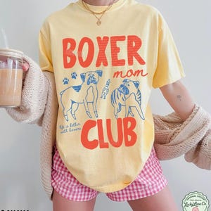 May include: A pale yellow short-sleeve t-shirt featuring the red text BOXER mom CLUB and blue line art illustrations of two Boxer dogs and a small toy. The shirt includes the blue script text Life is better with boxers and paw prints.
