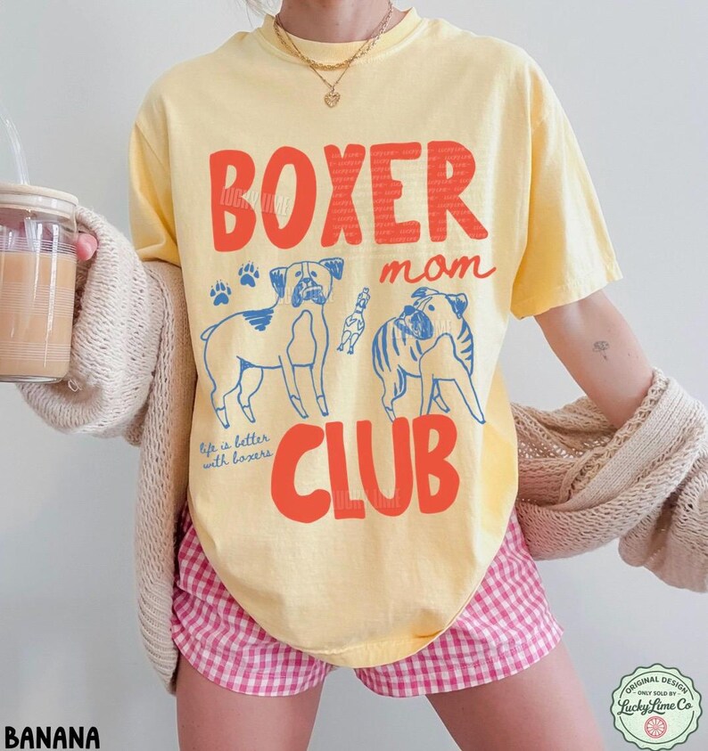 May include: A pale yellow short-sleeve t-shirt featuring the red text BOXER mom CLUB and blue line art illustrations of two Boxer dogs and a small toy. The shirt includes the blue script text Life is better with boxers and paw prints.