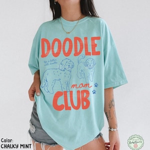Puede incluir: Una camiseta azul claro con las palabras "DOODLE MOM CLUB" en naranja. La camiseta presenta ilustraciones de perros doodle y la frase "life is better with doodles". El color de la camiseta es "CHALKY MINT".