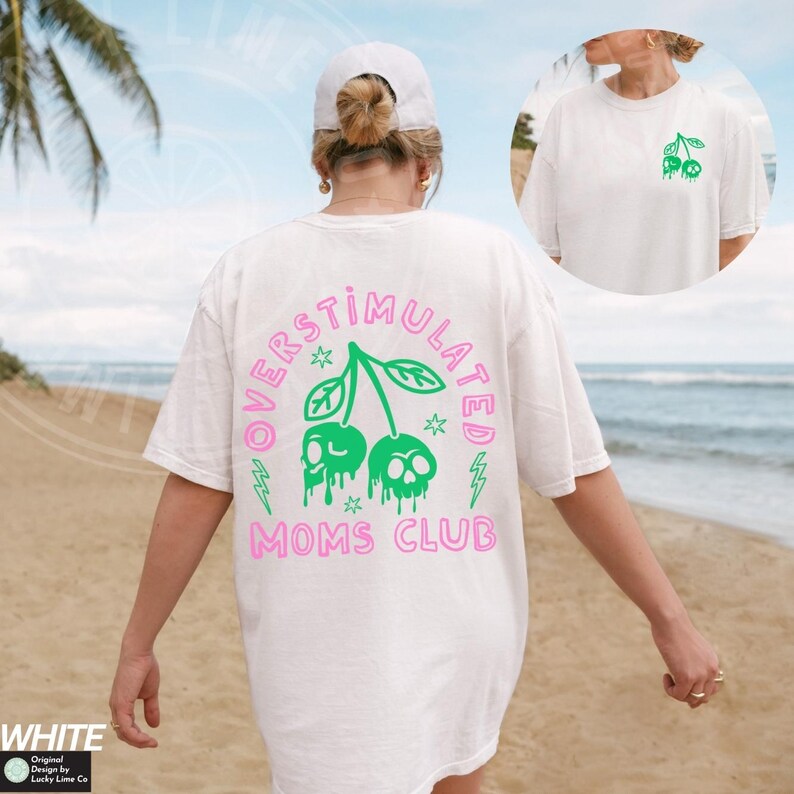 May include: White t-shirt featuring a pink and green graphic of two cherries with skulls on them. The text "Overstimulated Moms Club" is written in pink around the cherries.