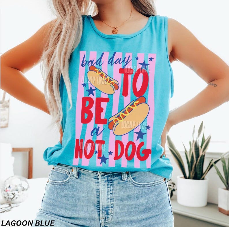 Op de afbeelding: Lagune blau tanktop met de tekst "bad day to be a hot dog" in rood en wit. Het ontwerp bevat hotdogs, blauwe sterren en roze en blauwe strepen. Een casual zomertop.