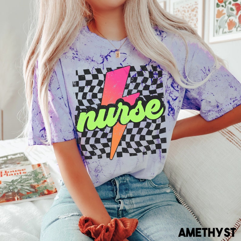 May include: A purple tie-dye t-shirt with a black and white chequered pattern and a lightning bolt graphic. The lightning bolt is pink, orange, and yellow. The word "nurse" is written in green letters on the lightning bolt.