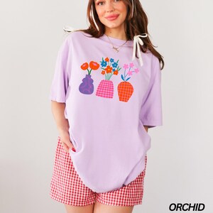 May include: A lavender t-shirt with a graphic of three vases with colorful flowers. The vases are different shapes and colors. The t-shirt is oversized and has a relaxed fit.
