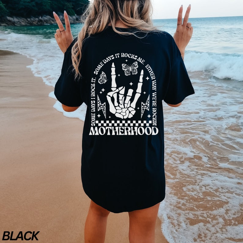 May include: Black t-shirt with a white graphic on the back. The graphic includes a skeleton hand making a rock and roll gesture, butterflies, and the text "SOME DAYS IT ROCKS ME. EITHER WAY WE'RE ROCKIN' MOTHERHOOD."