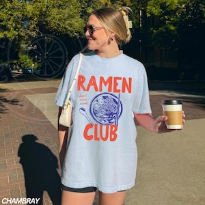 May include: A light blue t-shirt with the text "Ramen Club" in red and blue. The design features a bowl of ramen noodles with chopsticks and the text "Send more noodles".