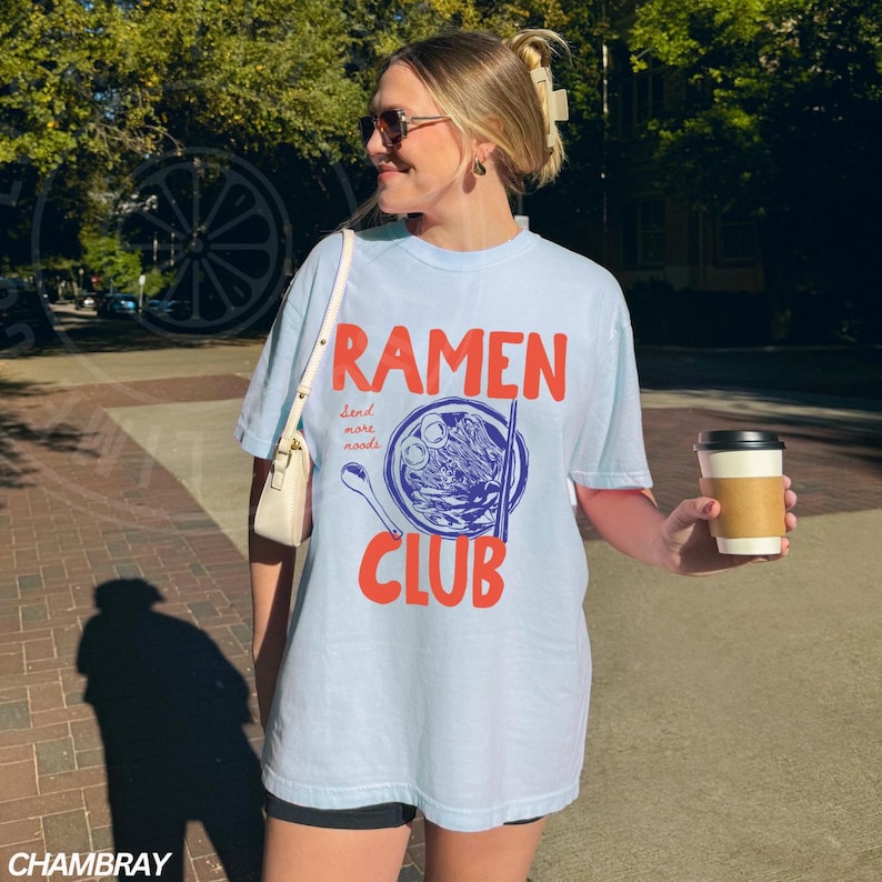 May include: A light blue t-shirt with the text "Ramen Club" in red and blue. The design features a bowl of ramen noodles with chopsticks and the text "Send more noodles".
