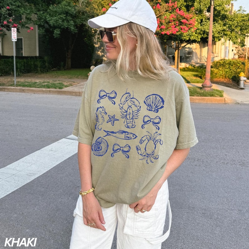 May include: A khaki green t-shirt with a blue graphic print of various sea creatures, including a seahorse, a lobster, a fish, a seashell, and a crab. The shirt has a relaxed fit and a crew neck.
