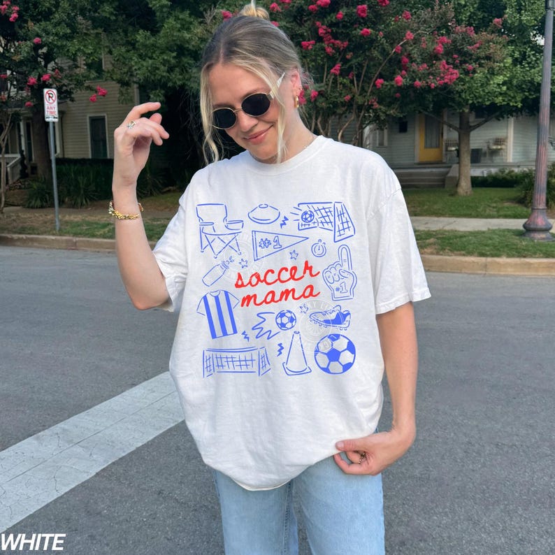 May include: White t-shirt with a blue and red graphic design featuring soccer-related illustrations and the text "soccer mama." The shirt has a relaxed fit and short sleeves. The word "WHITE" is printed at the bottom of the image.