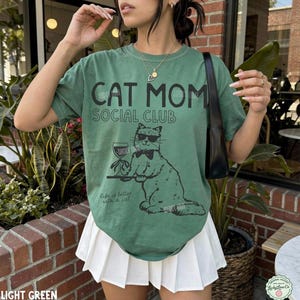 K&ouml;nnte beinhalten: Ein salbeigr&uuml;nes Kurzarm-T-Shirt mit dem schwarzen Schriftzug CAT MOM SOCIAL CLUB und einer Grafik einer Katze mit Sonnenbrille und Fliege, die ein Weinglas h&auml;lt. Darunter steht der Text Life is better with a cat in schwarzer Schrift.