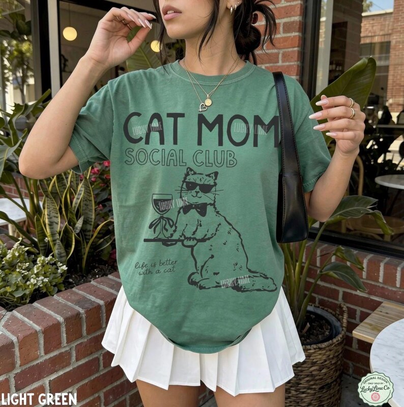K&ouml;nnte beinhalten: Ein salbeigr&uuml;nes Kurzarm-T-Shirt mit dem schwarzen Schriftzug CAT MOM SOCIAL CLUB und einer Grafik einer Katze mit Sonnenbrille und Fliege, die ein Weinglas h&auml;lt. Darunter steht der Text Life is better with a cat in schwarzer Schrift.