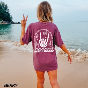 May include: Berry-colored t-shirt with a white graphic on the back. The graphic features a skeleton hand in a rock-and-roll gesture, butterflies, lightning bolts, and the word "MOTHERHOOD". Text on the shirt reads "SOME DAYS I ROCK IT. SOME DAYS IT ROCKS ME. EITHER WAY WE'RE ROCKIN".
