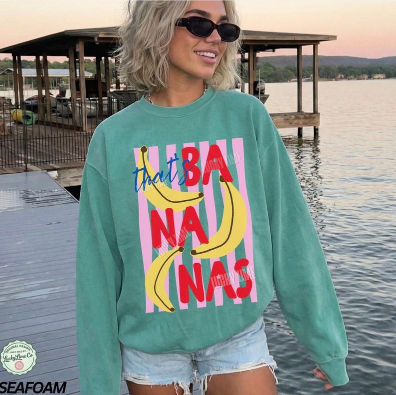 May include: Seafoam green sweatshirt with a graphic of bananas and the text "that's bananas" in a playful font. The design features yellow bananas and red text on a pink striped background. The sweatshirt is a casual style.