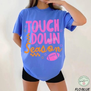 May include: A blue t-shirt with the words "TOUCH DOWN SEASON" in pink and orange, along with a pink football graphic. The shirt also has the text "LUCKY LIME" in pink. The shirt is a Flo Blue color.